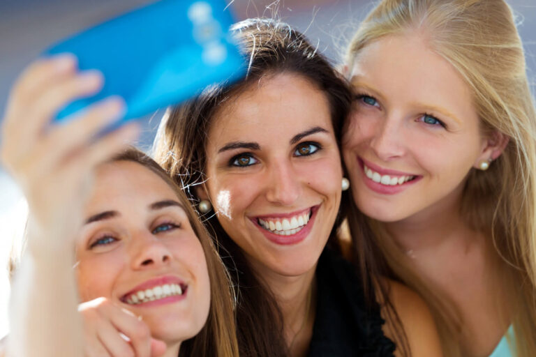 Três amigas sorrindo radiantes enquanto tiram uma selfie. Descubra se a prótese protocolo de zircônia no Brooklin é a ideal.